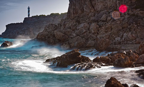Rough waves hit the rocks with a lighthouse in the background on a Balearics yacht charter